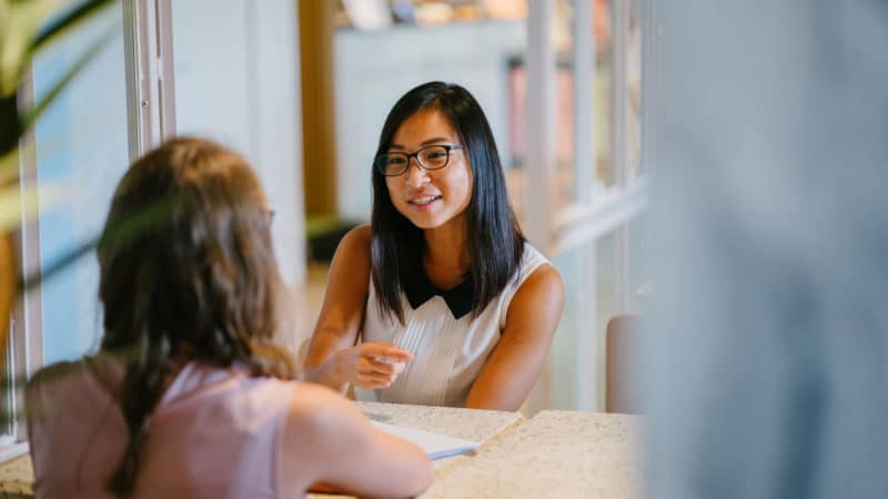 ```json
{
"alt": "Two women having a conversation at a table in a well-lit indoor setting.",
"caption": "Engaged in a lively discussion, two women share ideas over a cozy cafe table, creating connections and exchanging thoughts.",
"description": "Inside a bright and inviting room, two women are seated at a table, deep in conversation. One woman, wearing glasses and a sleeveless top, speaks animatedly while the other listens attentively. The setting suggests a warm, welcoming atmosphere with soft lighting and a comfortable environment, ideal for meaningful interactions. Keywords: conversation, women, indoor, discussion, connection."
}
```