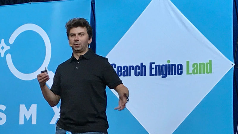 ```json
{
"alt": "Man in black polo presenting at a conference with Search Engine Land logo in the background.",
"caption": "Engaging presentation at a conference, set against a vibrant Search Engine Land backdrop.",
"description": "A speaker in a black polo shirt passionately gives a presentation at a conference. The backdrop features the Search Engine Land logo and is filled with shades of blue. The image captures a moment of engagement and interaction, ideal for SEO or digital marketing themes."
}
```