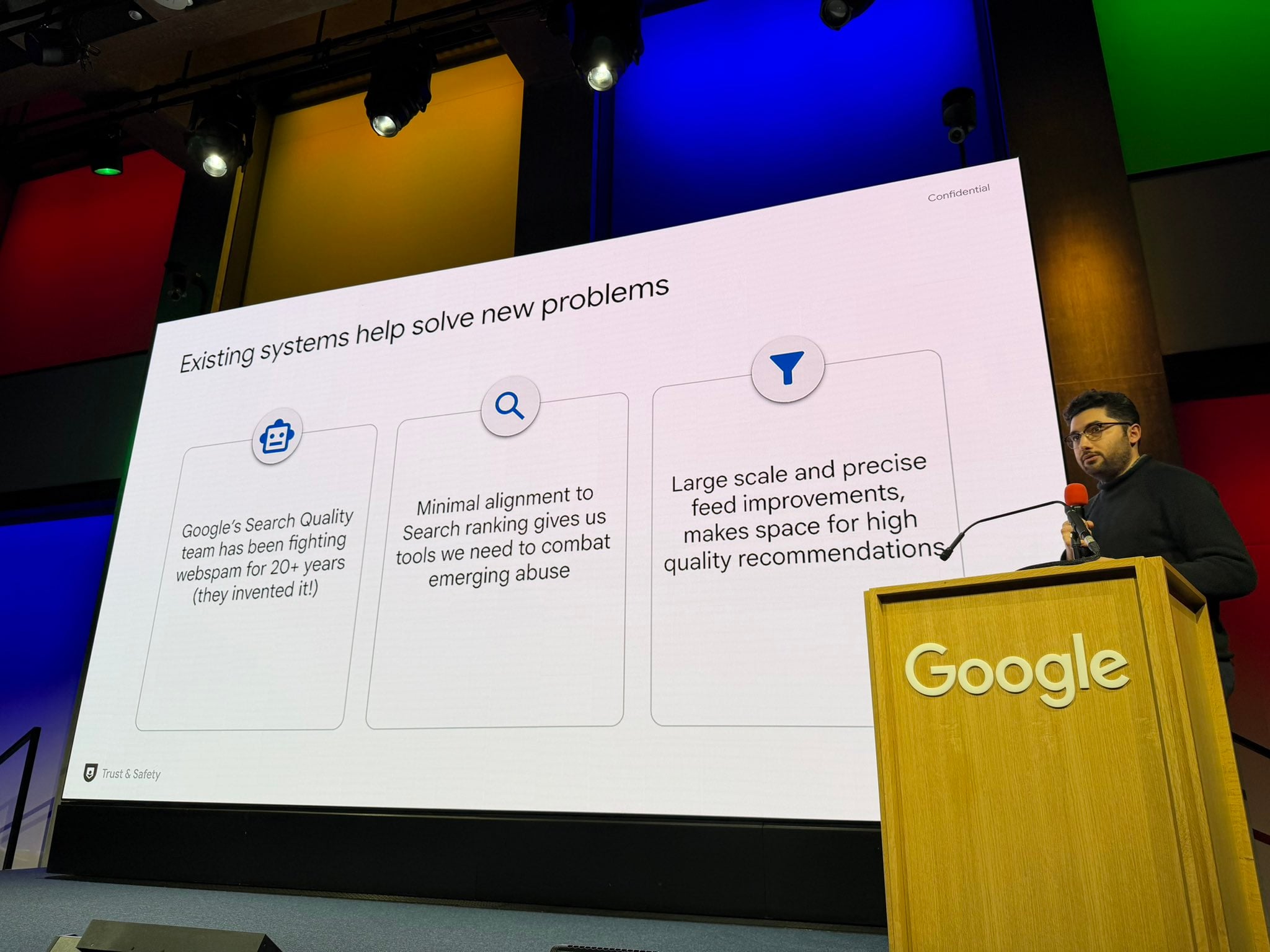```json
{
  "alt": "Person presenting at a Google event about search quality systems on a stage with colorful lights.",
  "caption": "A speaker at a Google event discusses solutions in combating web spam and enhancing search recommendations.",
  "description": "The image shows a speaker presenting at a Google event, standing at a wooden podium with a red microphone. On the large screen, there's information about Google's efforts to combat web spam through its search quality systems. The background features a colorful light display, emphasizing Google's innovative environment. Keywords: Google, presentation, search quality, web spam, technology event."
}
```