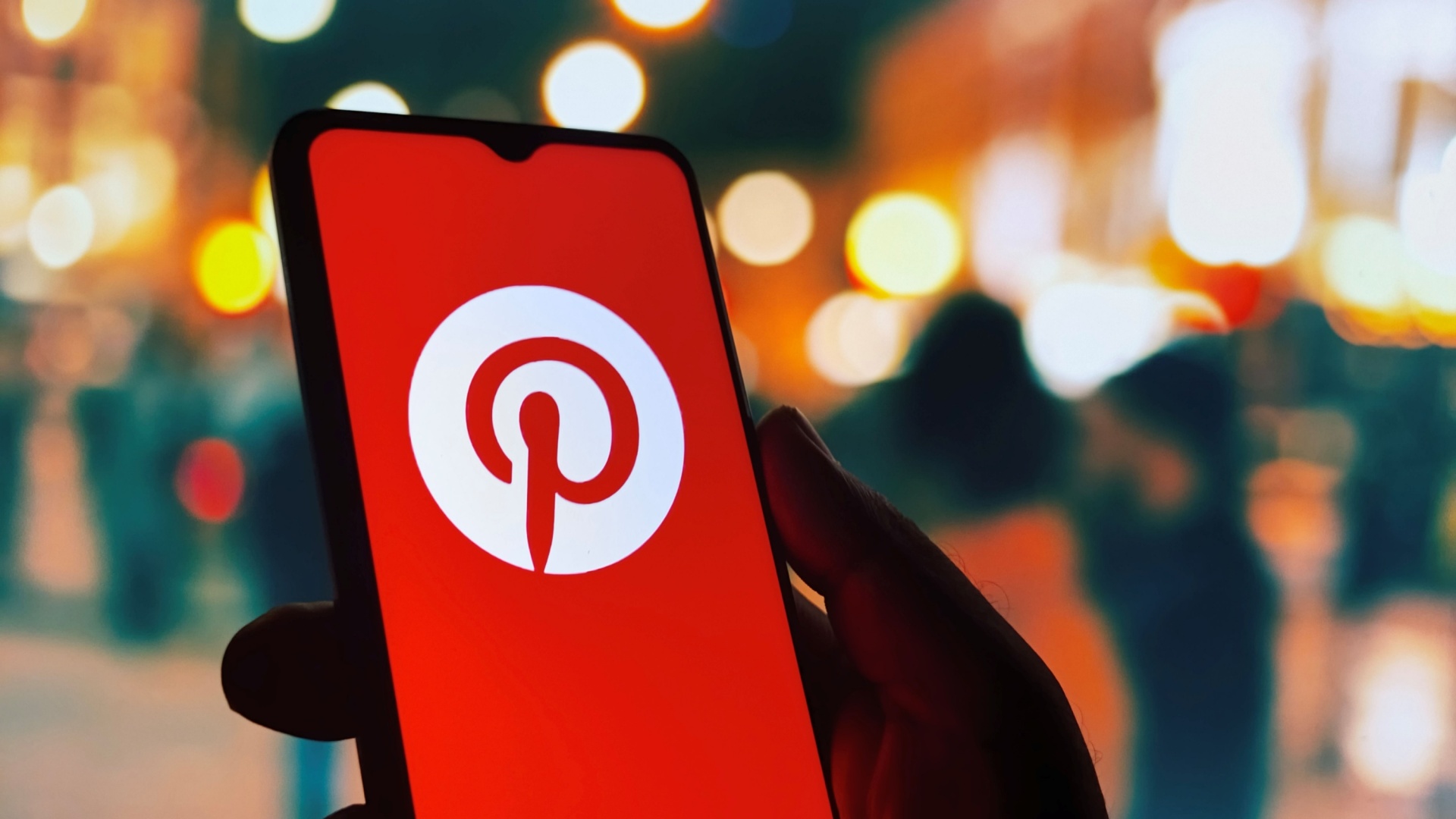 ```json
{
  "alt": "Smartphone displaying Pinterest app logo against a blurred city night background.",
  "caption": "Exploring creative ideas on Pinterest while the city lights sparkle in the background.",
  "description": "A silhouette of a hand holding a smartphone displaying the Pinterest app logo on its screen. The background reveals a beautifully blurred city night scene, with colorful bokeh lights creating an atmosphere of urban dynamism. This image captures the intersection of technology and creativity, highlighting the Pinterest app as a hub for inspiration amidst the vibrant city life."
}
```
