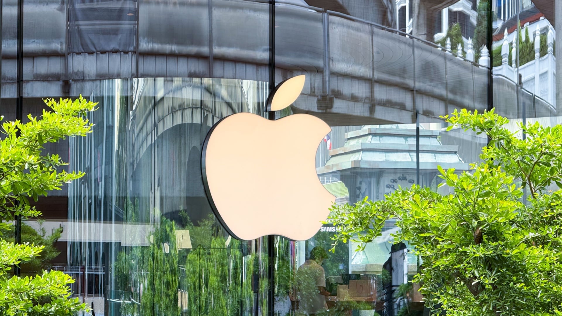 ```json
{
  "alt": "Apple logo on a glass building facade surrounded by green foliage.",
  "caption": "A striking Apple logo adorns the glass facade of a modern building, enhanced by lush green plants.",
  "description": "The image showcases the iconic Apple logo prominently displayed on a sleek glass building facade. Reflective surfaces capture the urban landscape, creating a dynamic visual interplay. Green foliage surrounds the area, adding a touch of nature to the modern architecture. This scene highlights the blend of technology and nature, symbolizing innovation and growth. Keywords: Apple, logo, building, glass facade, foliage, urban landscape."
}
```
