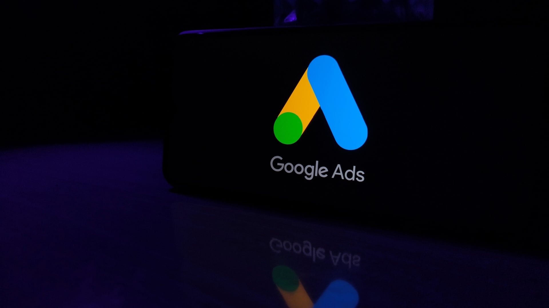 ```json
{
  "alt": "Google Ads logo on a dark background with reflection on surface.",
  "caption": "The vibrant Google Ads logo shines brightly, symbolizing digital marketing prowess, against a mysterious dark background.",
  "description": "This image showcases the Google Ads logo prominently displayed on a dark backdrop, with its colorful geometric design reflecting on a smooth surface. The contrast between the bright logo and the shadowy surroundings highlights the significance of Google Ads in the digital marketing landscape. Ideal for illustrating concepts related to online advertising, SEO, and digital marketing strategies."
}
```