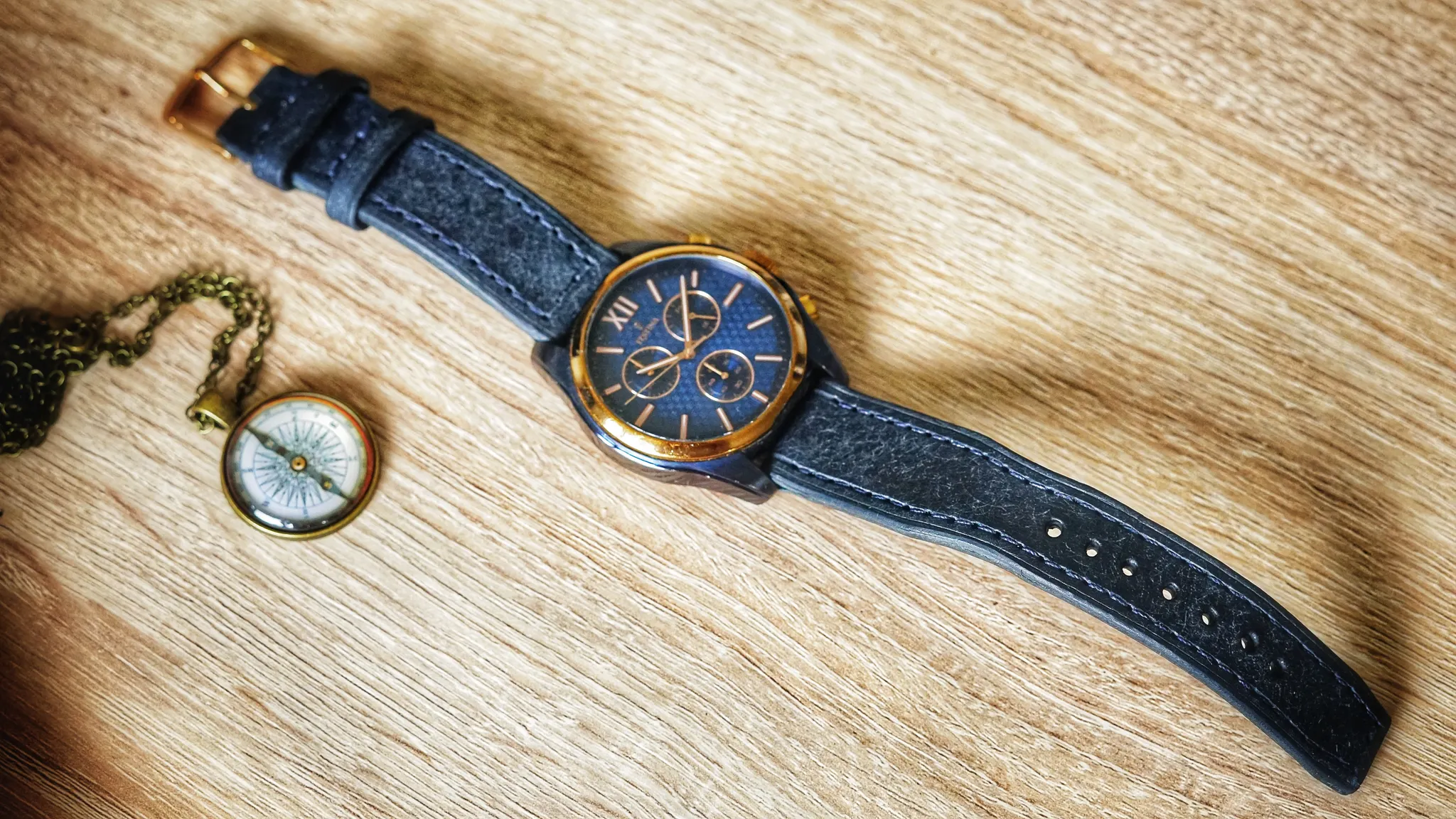 ```json
{
  "alt": "A wristwatch with a blue leather strap and a bronze casing lies next to a vintage brass compass on a wooden surface.",
  "caption": "Timeless elegance meets navigation with this stylish wristwatch and vintage brass compass duo, perfectly paired on a rustic wooden table.",
  "description": "The image features a sophisticated wristwatch with a blue leather strap and a bronze casing set atop a wooden surface. Next to it lies a vintage brass compass with an intricate chain, creating a harmonious blend of style and exploration. The rich textures and warm tones of the wood enhance the elegance of both pieces, making this a perfect symbol of timeless grace and adventure. Keywords: wristwatch, compass, leather strap, bronze casing, vintage, elegance."
}
```