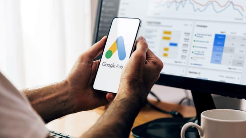 ```json
{
  "alt": "Person holding a smartphone with Google Ads logo, computer screen displaying analytics data in background.",
  "caption": "Digital marketing on the go: Navigating Google Ads analytics seamlessly between mobile and desktop.",
  "description": "The image shows a person holding a smartphone displaying the Google Ads logo, indicating mobile accessibility of the platform. In the background, a computer monitor shows detailed analytics and data charts related to advertising campaigns. This setup highlights the integration of digital marketing tools across devices, emphasizing ease of access and analysis in work environments focused on online advertising. Key elements include digital marketing, analytics, Google Ads, and mobile integration."
}
```