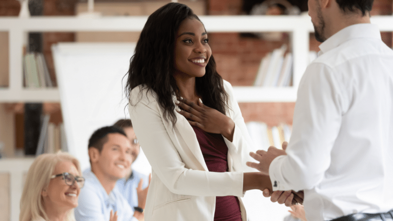 ```json
{
  "alt": "Smiling woman in a white blazer shaking hands with a man in an office setting, others clapping in the background.",
  "caption": "A joyful moment of congratulations as colleagues applaud the woman in the white blazer during a professional exchange.",
  "description": "In a lively office setting, a woman wearing a white blazer joyfully shakes hands with a man. The background features clapping colleagues, indicating a celebratory or congratulatory event. The scene suggests a professional environment, possibly during a meeting or presentation. Keywords: handshake, office, celebration, colleagues, professional setting."
}
```