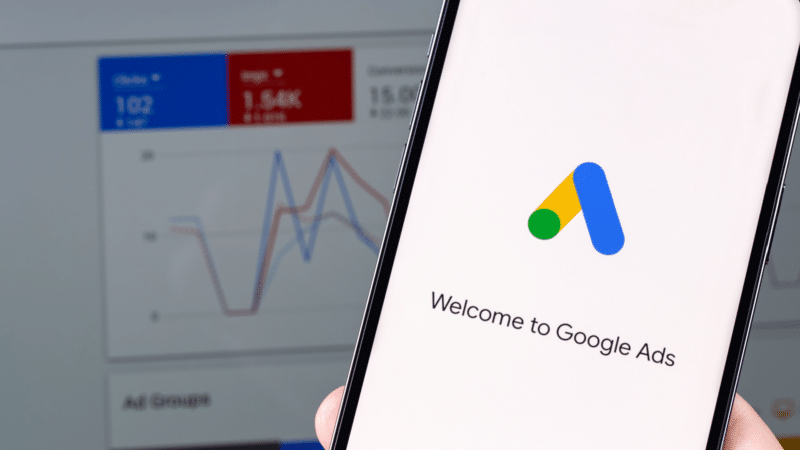 ```json
{
  "alt": "Smartphone displaying Google Ads logo with analytics chart in the background.",
  "caption": "Dive into digital marketing with Google Ads! A smartphone showcases the iconic logo in front of analytics, signaling growth and opportunity.",
  "description": "The image features a smartphone showing the Google Ads logo and 'Welcome to Google Ads' message. In the background, a blurred screen displays an analytics chart, depicting data trends in blue and red lines. This setup represents digital marketing strategies and the use of analytics in optimizing ad performance, ideal for illustrating topics related to SEO, online advertising, and data analysis."
}
```