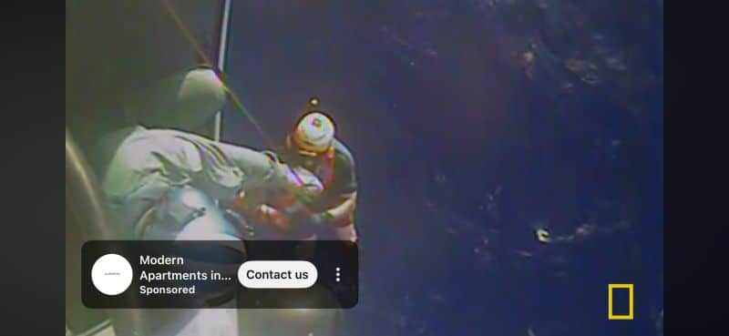 ```json
{
  "alt": "Rescue worker assisting a person in distress over a body of water, with a National Geographic logo and advertisement overlay.",
  "caption": "A dramatic rescue operation unfolds over the deep blue sea, capturing a heroic moment against the backdrop of vast ocean waves.",
  "description": "This image shows a rescue worker aiding a person in distress, suspended above an expansive ocean. The scene is intense, highlighting the bravery and urgency of rescue operations at sea. The image includes a National Geographic logo, suggesting it's part of a related documentary or news feature. An advertisement banner is visible, offering modern apartment details, adding a layer of commercial context to the dramatic scene."
}
```