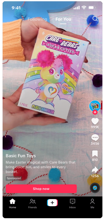 ```json
{
  "alt": "Hand holding a Care Bears Sugar Cuties toy box, featuring a colorful bear with a rainbow background.",
  "caption": "Brighten up your Easter with Care Bears Sugar Cuties! This colorful scented plush dazzles with a rainbow vibe, perfect for any festive basket.",
  "description": "The image displays a hand holding a Care Bears Sugar Cuties toy box. The design features a vibrant, colorful bear character against a rainbow background. This toy is highlighted as scented and is described as a 'Plush Dangler'. The screen is part of a TikTok video, featuring various engagement icons on the side and a 'Shop now' button below. This playful presentation is part of a sponsored promotion by Basic Fun Toys, aiming to add color and joy to Easter celebrations. Keywords: Care Bears, Sugar Cuties, toy, Easter, rainbow, plush, scent, Basic Fun Toys."
}
```