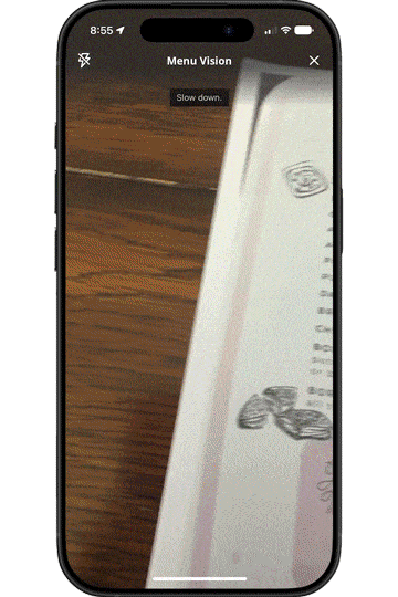 ```json
{
"alt": "Smartphone app scanning a document, displaying a 'Slow down' prompt.",
"caption": "Experience the efficiency of document scanning with this smartphone app, guiding users with a 'Slow down' prompt for optimal results.",
"description": "This image shows a smartphone screen displaying an app named 'Menu Vision' being used to scan a document. The on-screen prompt 'Slow down' appears, indicating the app's suggestion to move the phone slower for better scanning accuracy. The background displays a wooden surface, and part of the document includes text and drawings. This scene highlights the app's interactive and user-friendly features, which enhance document scanning efficiency."
}
```