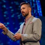 ```json
{
  "alt": "Person speaking on stage, gesturing, in a gray suit with a blue backdrop.",
  "caption": "Engaged in thought-provoking discourse, the speaker captivates the audience under a striking blue backdrop.",
  "description": "A person stands on a stage, speaking and gesturing confidently. They are dressed in a gray suit and light checkered shirt, with a blue background that adds depth to the scene. The image captures the essence of a dynamic presentation, showcasing public speaking in a professional setting. Keywords: public speaking, presentation, keynote, professional speaker, stage presentation."
}
```