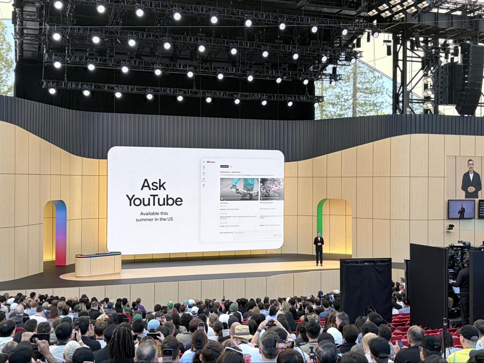 ```json
{
  "alt": "Large tech presentation with speaker on stage, introducing 'Ask YouTube', to a full audience.",
  "caption": "Innovation takes the stage as 'Ask YouTube' is unveiled in front of an excited crowd, promising new features available this summer.",
  "description": "A large tech event features a speaker presenting 'Ask YouTube' on a massive screen, announcing its availability in the U.S. this summer. The venue is packed with attendees capturing the moment on their devices. Stage lighting and modern design elements underscore the futuristic theme of the event. This announcement is part of a wider tech conference, drawing a diverse crowd eager for new advancements. Keywords: tech presentation, Ask YouTube, innovation, audience, event."
}
```