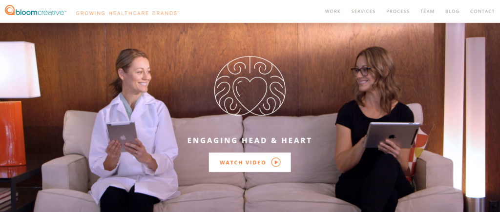 ```json
{
  "alt": "Two women sitting on a couch, engaging with tablets, in a professional setting with a 'Watch Video' button.",
  "caption": "Discover the synergy of intellect and emotion as two women engage on tablets, symbolizing the blend of 'Head & Heart' in healthcare branding.",
  "description": "This image features two women seated comfortably on a cream-colored sofa, each interacting with a tablet. The background has a warm wooden texture, and a graphic of a heart symbol divides the space, emphasizing a balance between emotional and thoughtful engagement. The women appear involved in a healthcare-related branding context, indicative of Bloom Creative’s focus. The image invites viewers to 'Watch Video,' suggesting further exploration of their innovative approach."
}
```