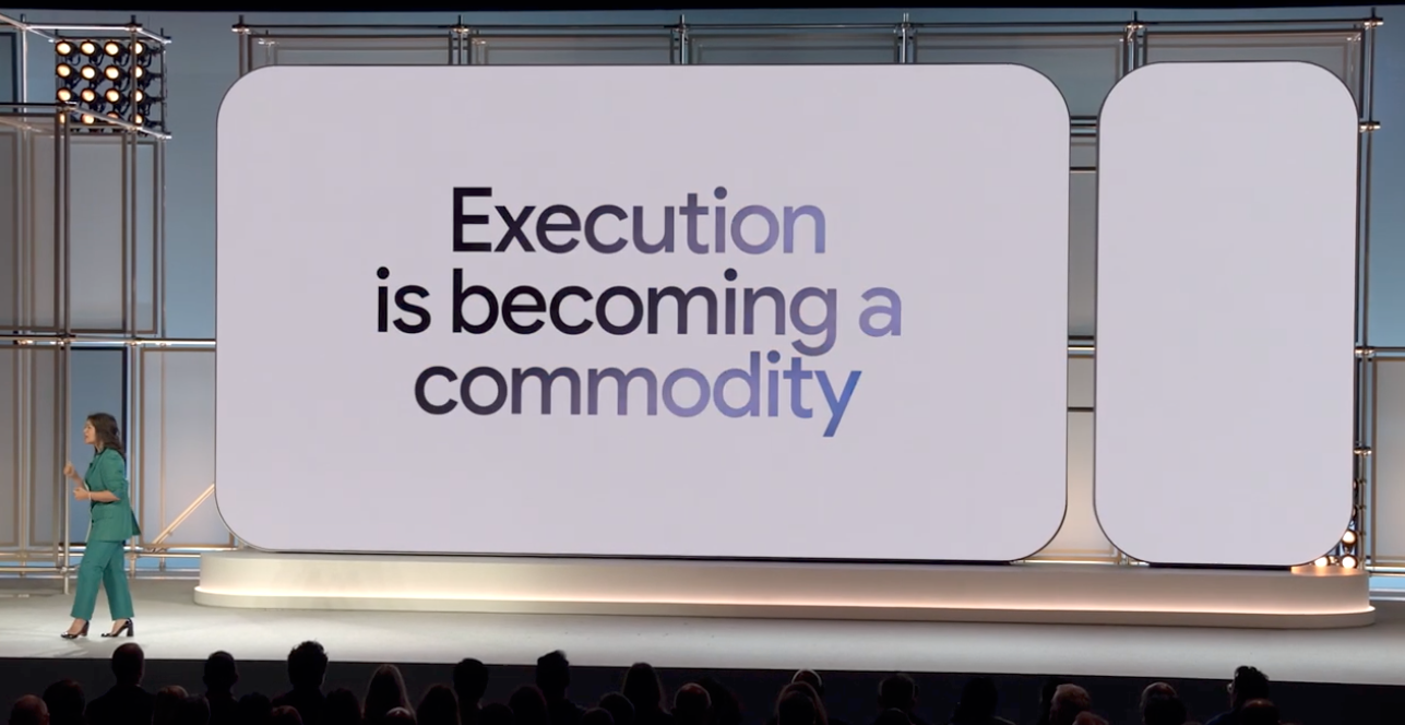 ```json
{
  "alt": "Speaker in teal suit on stage with large screen displaying 'Execution is becoming a commodity'.",
  "caption": "A thought-provoking message, 'Execution is becoming a commodity,' displayed on a large screen during an engaging talk.",
  "description": "The image depicts a speaker in a teal suit presenting on stage in front of a large audience. A prominent screen behind them showcases the phrase, 'Execution is becoming a commodity.' The scene suggests a conference or seminar setting, emphasizing the importance of innovative thinking in modern industries. The stage is well-lit with a metal framework and soft background lighting, providing a professional and focused atmosphere. Keywords: execution, commodity, presentation, innovation, conference."
}
```