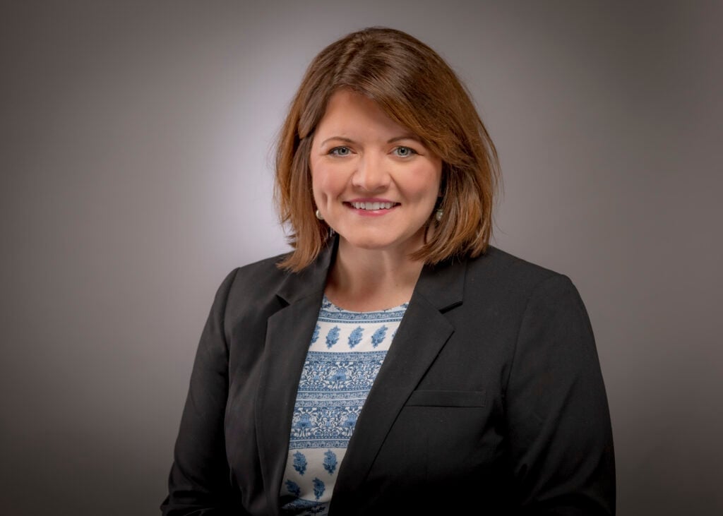 ```json
{
  "alt": "Smiling woman with short brown hair wearing a black blazer and patterned blouse against a gray background.",
  "caption": "A confident smile against a neutral backdrop. This portrait captures professionalism and warmth, featuring a woman in a black blazer and patterned blouse.",
  "description": "This image features a woman with short brown hair smiling warmly at the camera. She is wearing a black blazer over a blue and white patterned blouse. The neutral gray background adds a professional touch, making this portrait ideal for business or profile purposes. Keywords: portrait, woman, professional, blazer, smile."
}
```