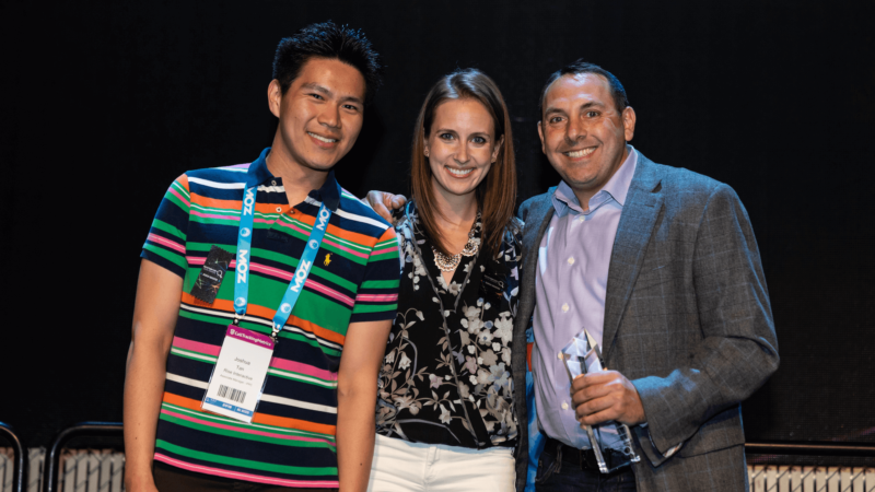 ```json
{
  "alt": "Three people smiling at an event, one holding an award.",
  "caption": "Celebrating Achievements: A trio beams with joy as one holds an award, capturing a moment of success and camaraderie.",
  "description": "In this image, three individuals are standing together at an event. The person on the left is wearing a colorful striped shirt and a lanyard with name tags. The person in the center is dressed in a black floral blouse and white pants, while the person on the right, dressed in a gray jacket over a light shirt, holds an award. They are all smiling, suggesting a celebratory occasion. The dark backdrop highlights their joyful expressions. Keywords: event, award, celebration, people, success."
}
```