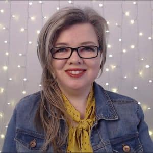 ```json
{
  "alt": "Smiling woman with glasses in denim jacket against a backdrop of string lights.",
  "caption": "A cheerful moment captured as she stands against a mesmerizing backdrop of twinkling string lights, blending casual style with a touch of glamour.",
  "description": "The image features a woman with glasses, smiling warmly while wearing a denim jacket and a yellow scarf. Behind her, a series of string lights create a cozy and festive atmosphere. The contrast between her casual attire and the glamorous lighting adds an engaging visual dynamic, perfect for themes of warmth, style, or celebration."
}
```