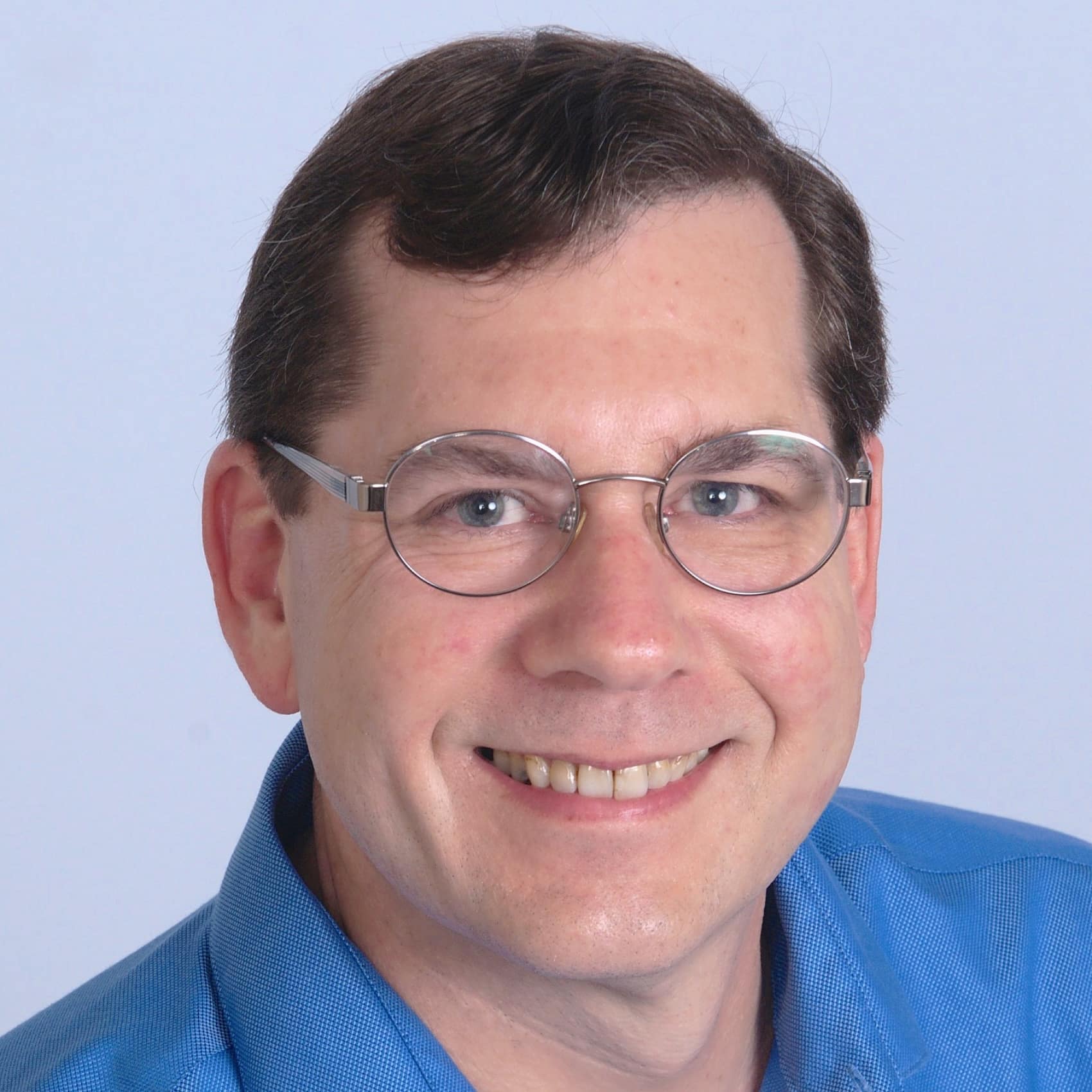 ```json
{
  "alt": "Portrait of a smiling man with glasses, wearing a blue shirt against a light background.",
  "caption": "A cheerful individual captured in a professional portrait, showcasing a warm smile and approachable demeanor.",
  "description": "This image depicts a close-up portrait of a smiling man wearing thin-rimmed glasses and a blue collared shirt. The backdrop is a simple light color, which enhances the subject's friendly and welcoming expression. The photograph is taken with good lighting, highlighting his facial features clearly, making it suitable for professional or personal use in profiles or presentations."
}
```