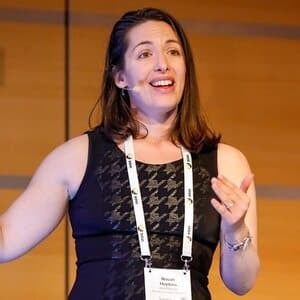 ```json
{
  "alt": "Woman in a black dress speaking at a conference with a microphone.",
  "caption": "Engaging and insightful, she captivates the audience during her dynamic conference presentation.",
  "description": "A woman wearing a black dress is speaking passionately at a conference. She is using a microphone attached to her face and gesturing with her hands, suggesting a lively presentation. The background features a wooden paneling typically found in professional or academic settings. Her conference badge suggests she is a keynote speaker or panelist. The image conveys a sense of professionalism and engagement, making it ideal for topics related to public speaking, leadership, or conferences."
}
```