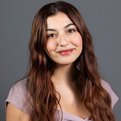 ```json
{
  "alt": "Portrait of a woman with long brown hair wearing a light purple top, smiling against a gray background.",
  "caption": "A warm smile and confident demeanor define this portrait, capturing the essence of positive energy against a neutral gray backdrop.",
  "description": "This image features a woman with long, wavy brown hair. She is wearing a light purple top and smiling gently at the camera, set against a smooth gray background. The soft lighting highlights her friendly expression, making this photo ideal for professional or casual contexts. It is perfect for use in profiles, articles, or media requiring a positive and approachable image."
}
```