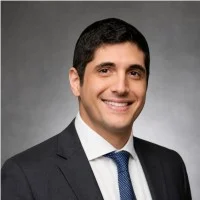 Smiling man in a dark suit, white shirt, and blue patterned tie against a gray studio background.