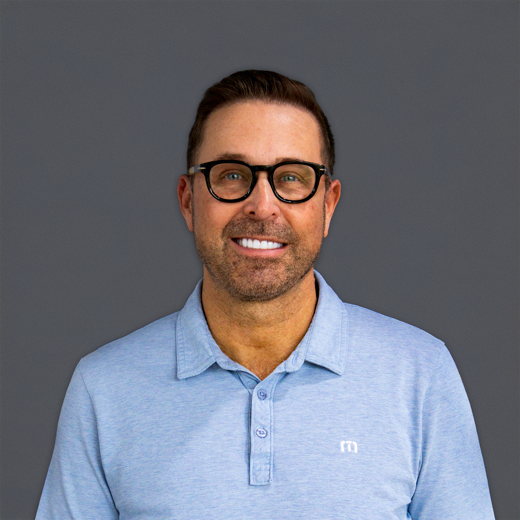 Smiling man with black-framed glasses and a light blue polo shirt against a dark gray background.